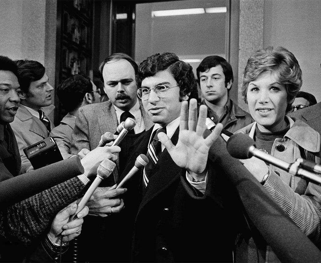 A a black and white archival photo from Watergate of a man holding up his hand to reporters.