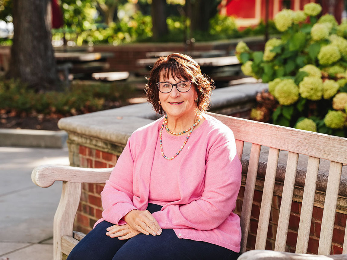 A woman in a pink sweater sits on a bench on a college campus