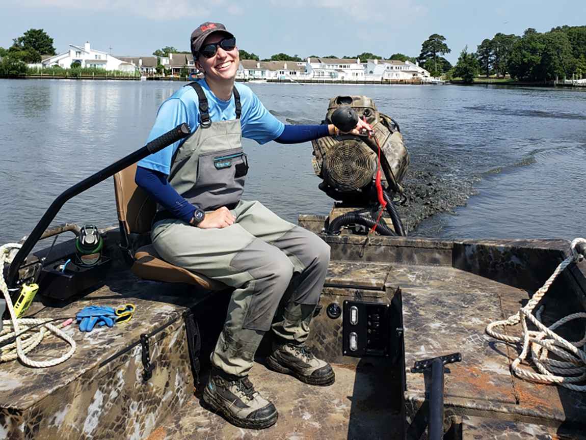 A young person sits on a boat wearing waders and boots