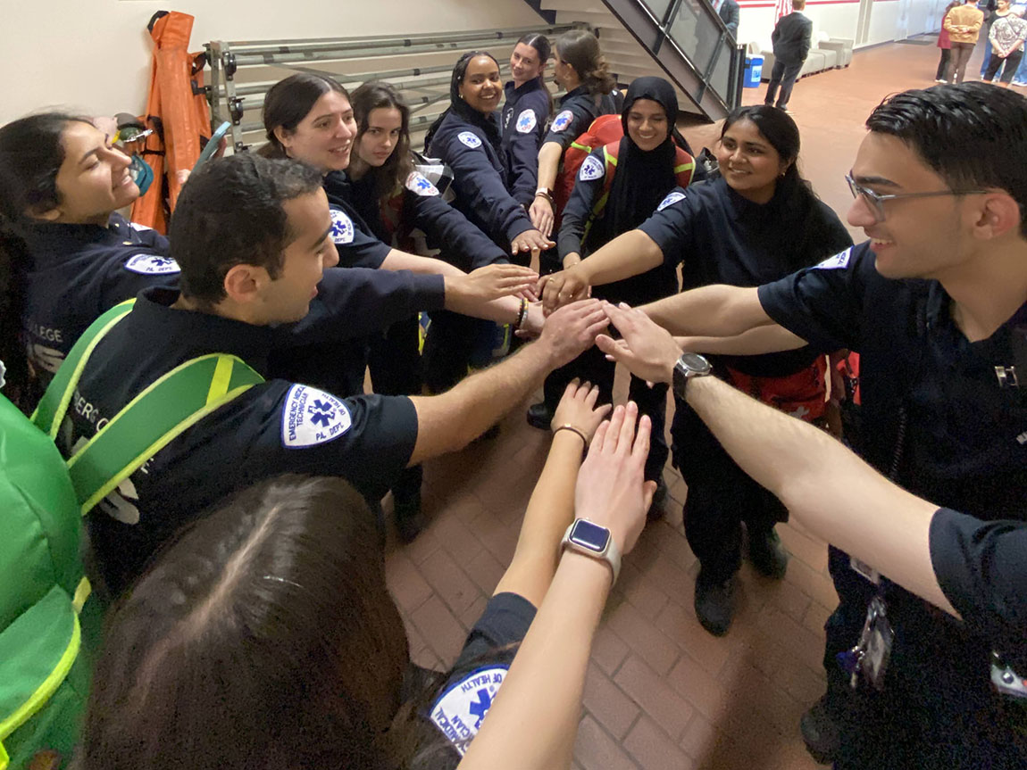 A group of college students in EMS uniforms put their hands into the middle of a circle