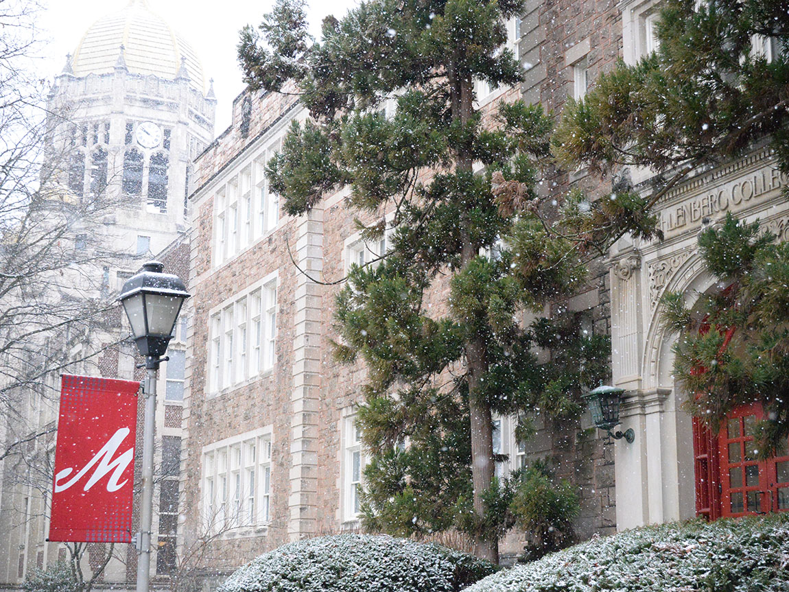 College buildings with snow falling in the foreground and a red flag with a white cursive M on it