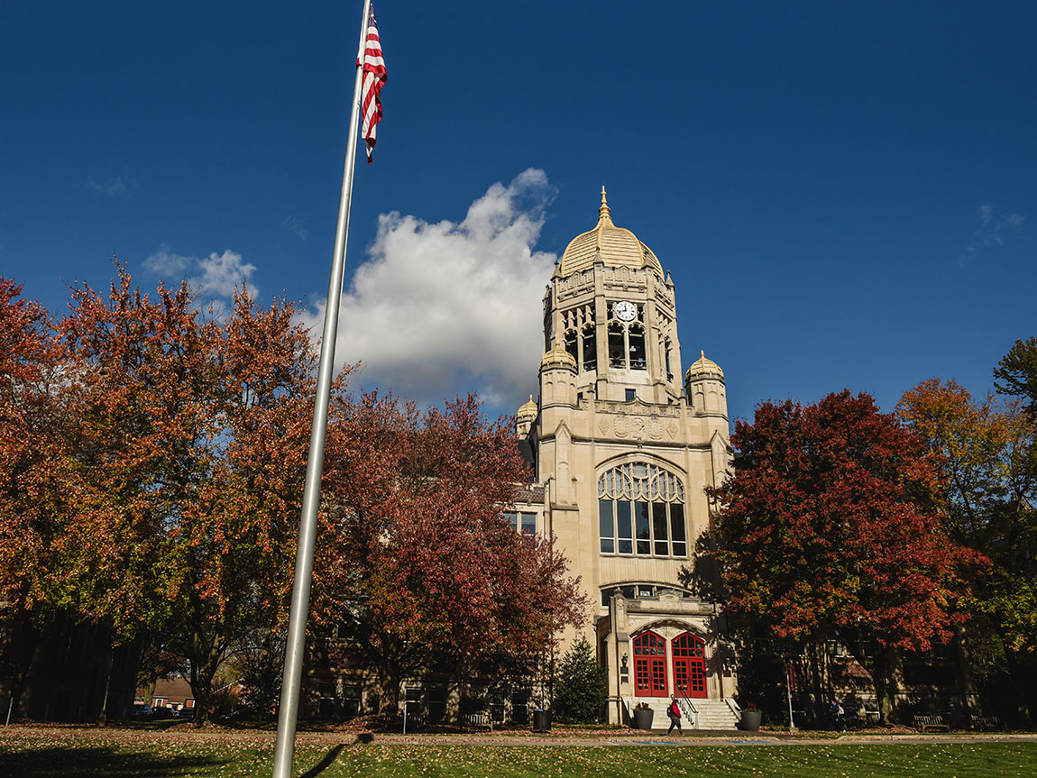 A large, old building topped with a bell tower with fall foliage and blue skies surrounding it