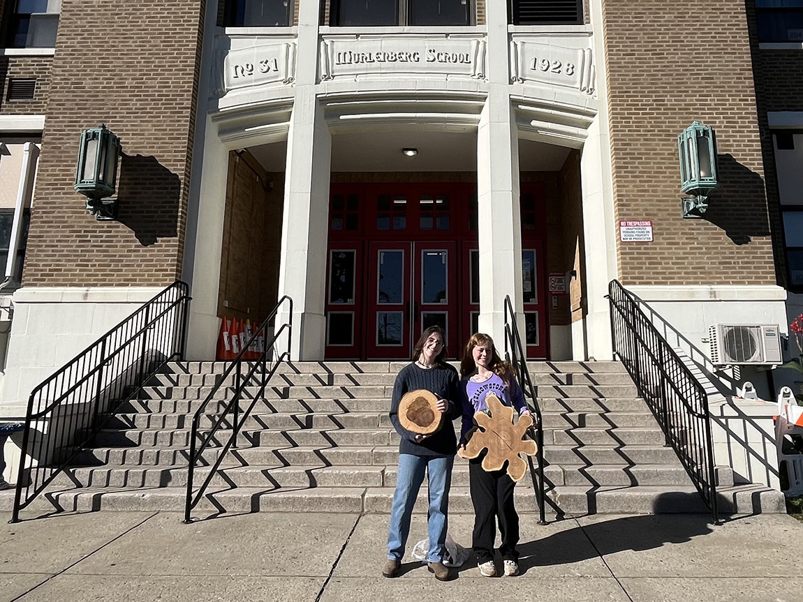 Two college students hold cross-sections of tree trunks outside an elementary school