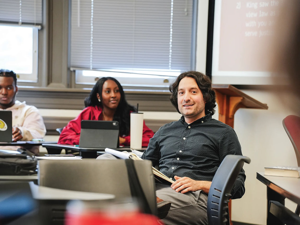 A college professor sits in a desk with students near him