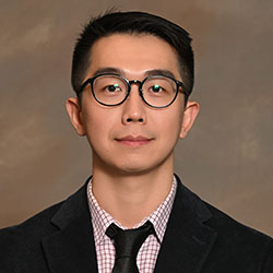 A headshot of a college professor with dark hair, glasses, and a black tie on