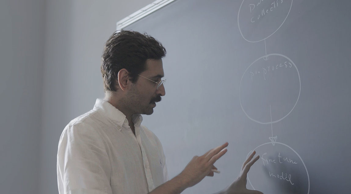 A college professor stands in front of a blackboard and points to it