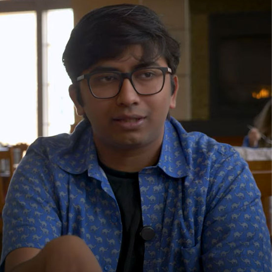 A college student in a blue shirt and glasses speaks to the camera while seated in a dining hall.