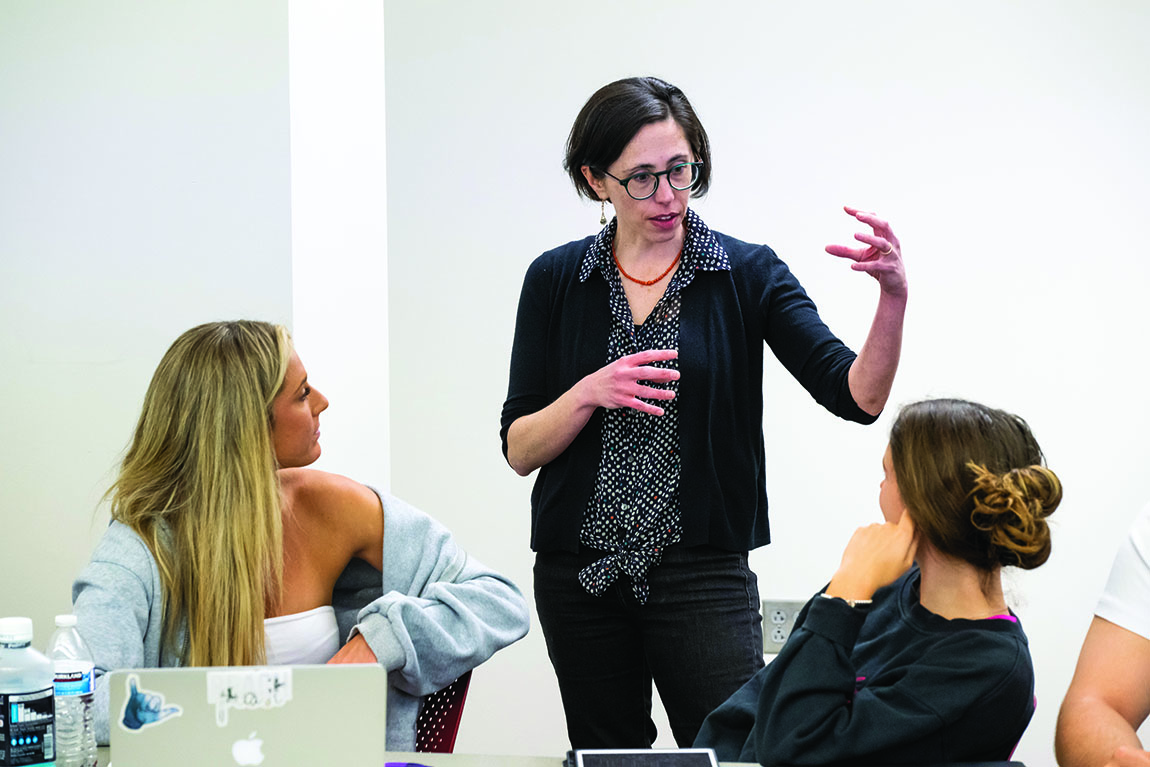 A standing professor gestures in a classroom as she speaks to two seated students