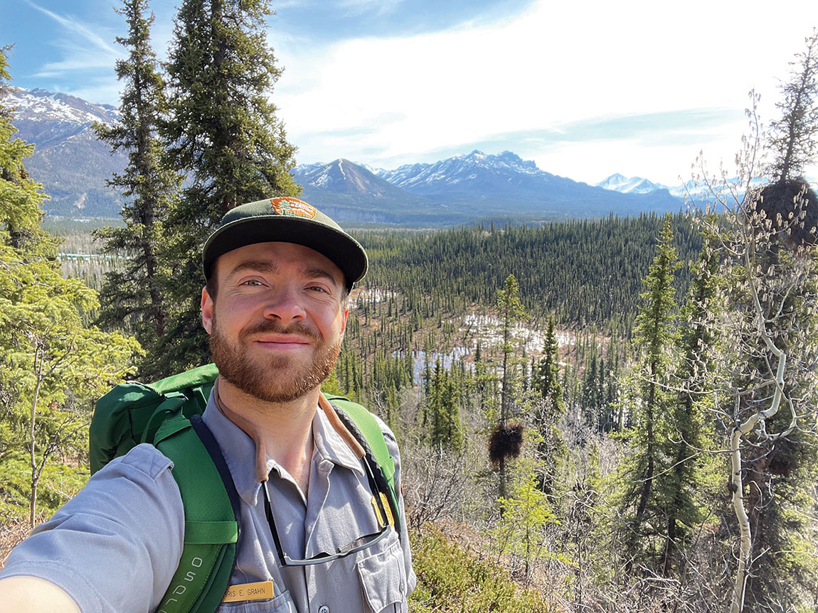 A young adult takes a selfie with snow-capped mountains and a pine forest behind him