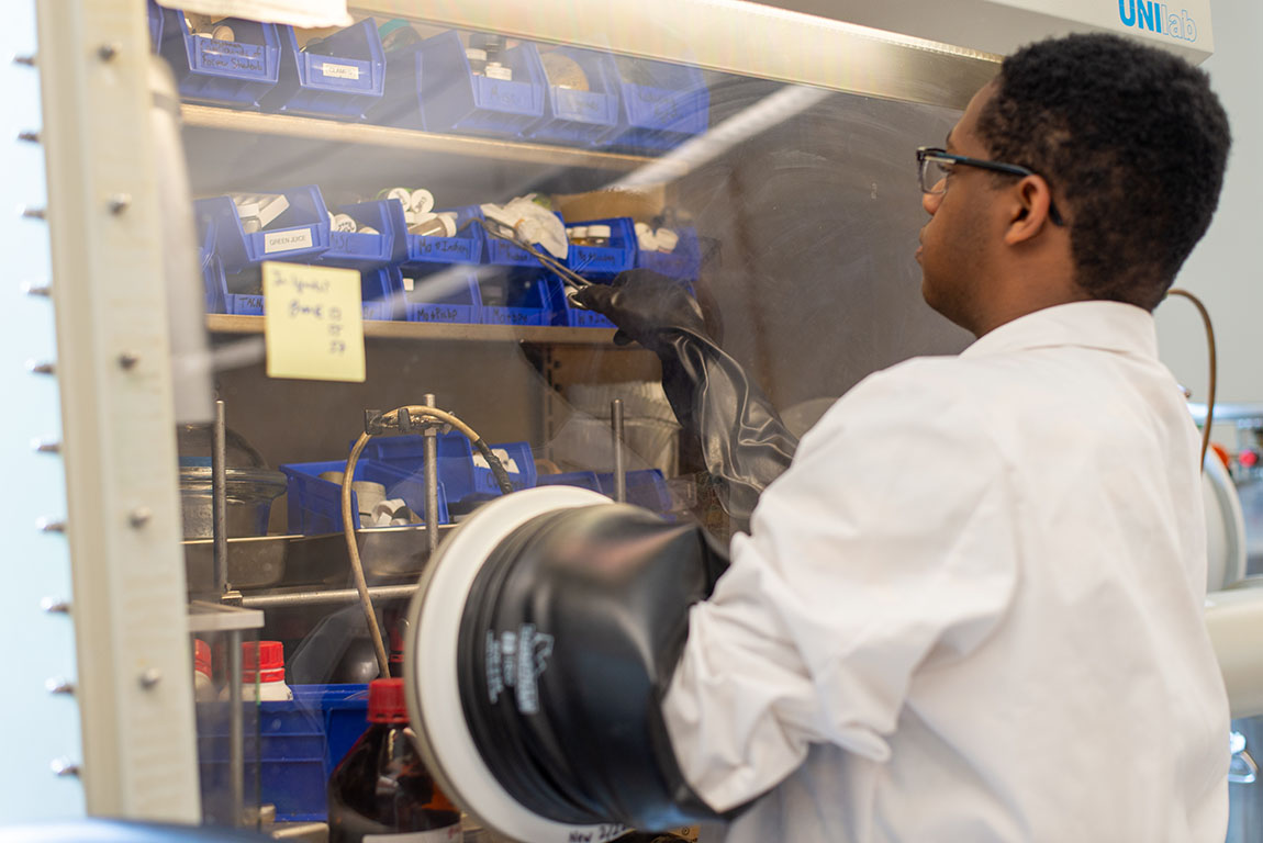 A college student in a white lab coat uses a glove box in a chemistry lab