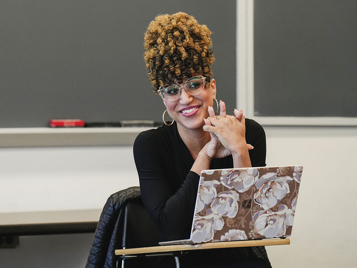 A college professor sits at a desk with her laptop in front of her and smiles at her students