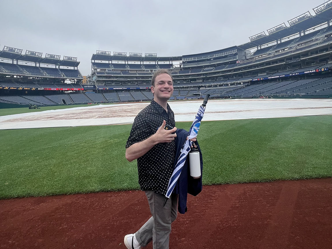 A college student gives a thumbs up to the camera while walking on a baseball field inside a large stadium