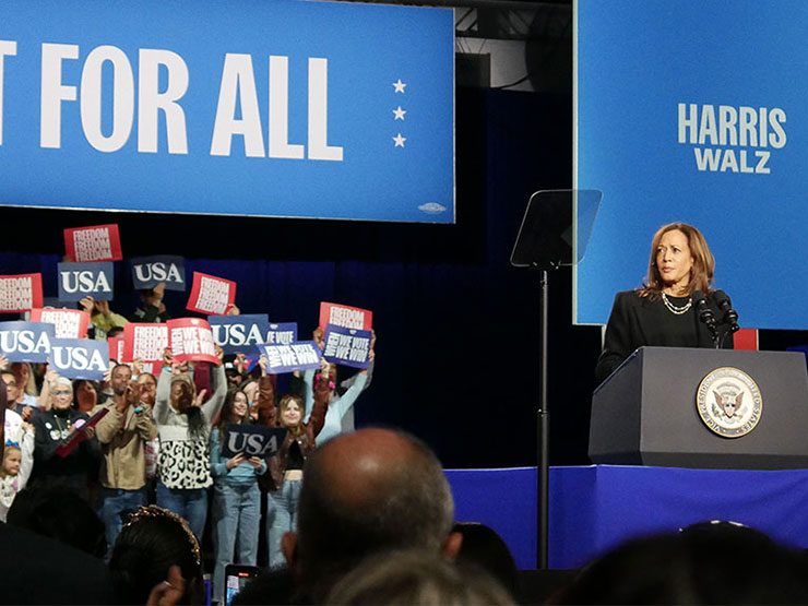 Vice President Kamala Harris stands at a podium with a crowd nearby holding campaign signs.