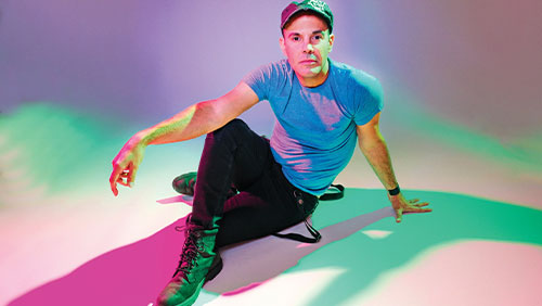 A man poses on the ground, hand over a bent knee, in a room with colorful lighting.