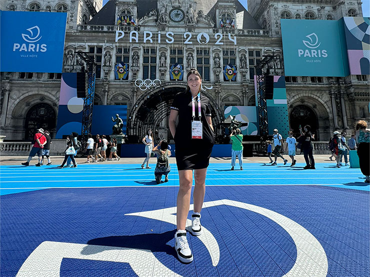 A smiling woman with a lanyard around her neck stands outside of gothic building decorated with signs for the Paris Olympics.