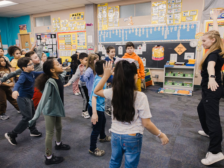 College students lead elementary-age students in dance inside a grade school classroom.