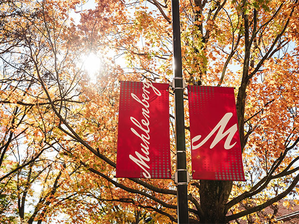 A pair of red Muhlenberg College banners hang from a lightpost against the backdrop of autumn foaliage.