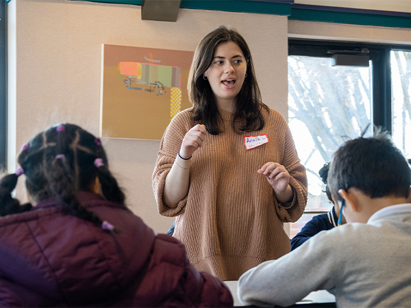 A college student wearing a name tag speaks to a group of elementary-age students in a classroom.